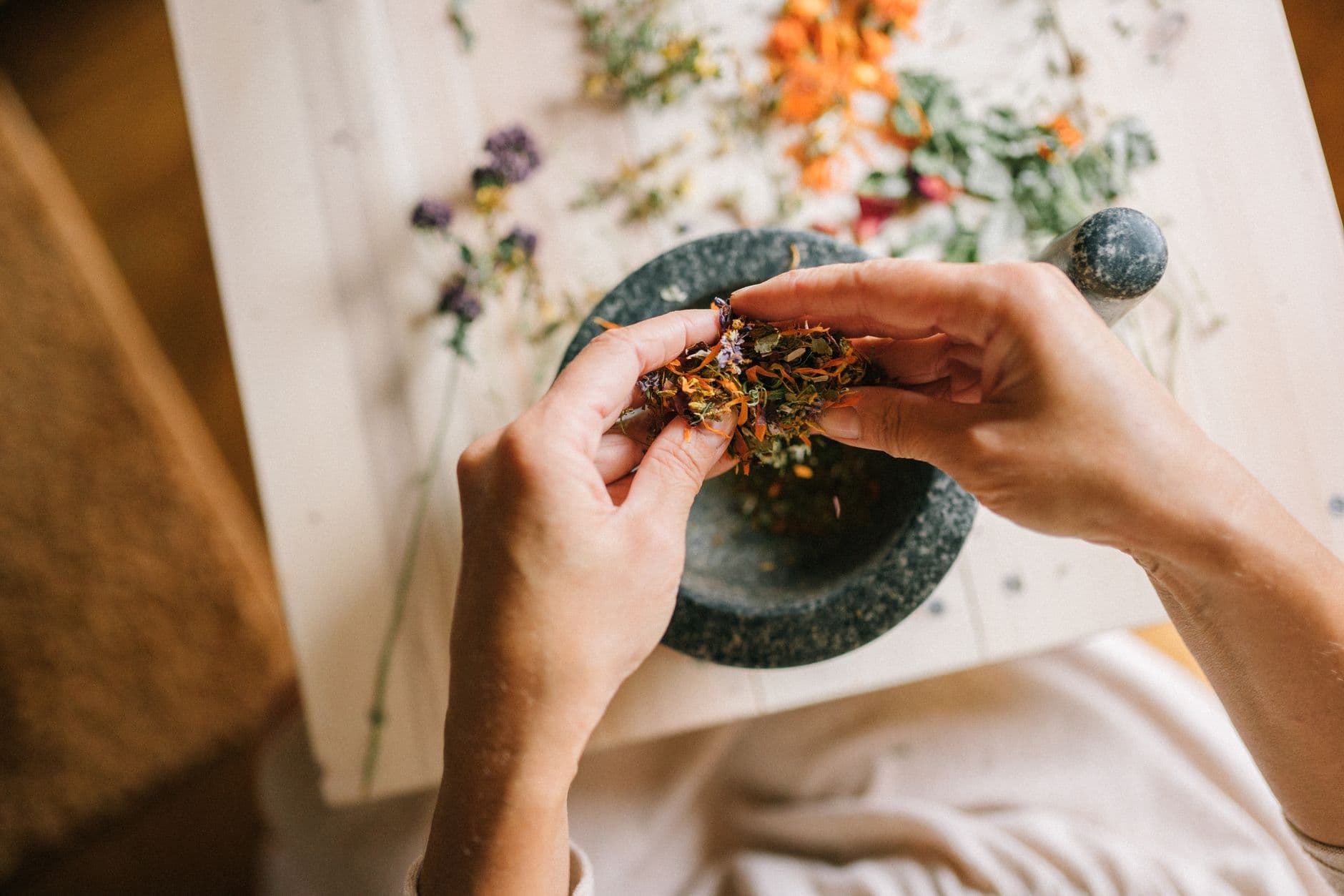 Hands working dried flowers and herbs together in a stone mortar on a bench