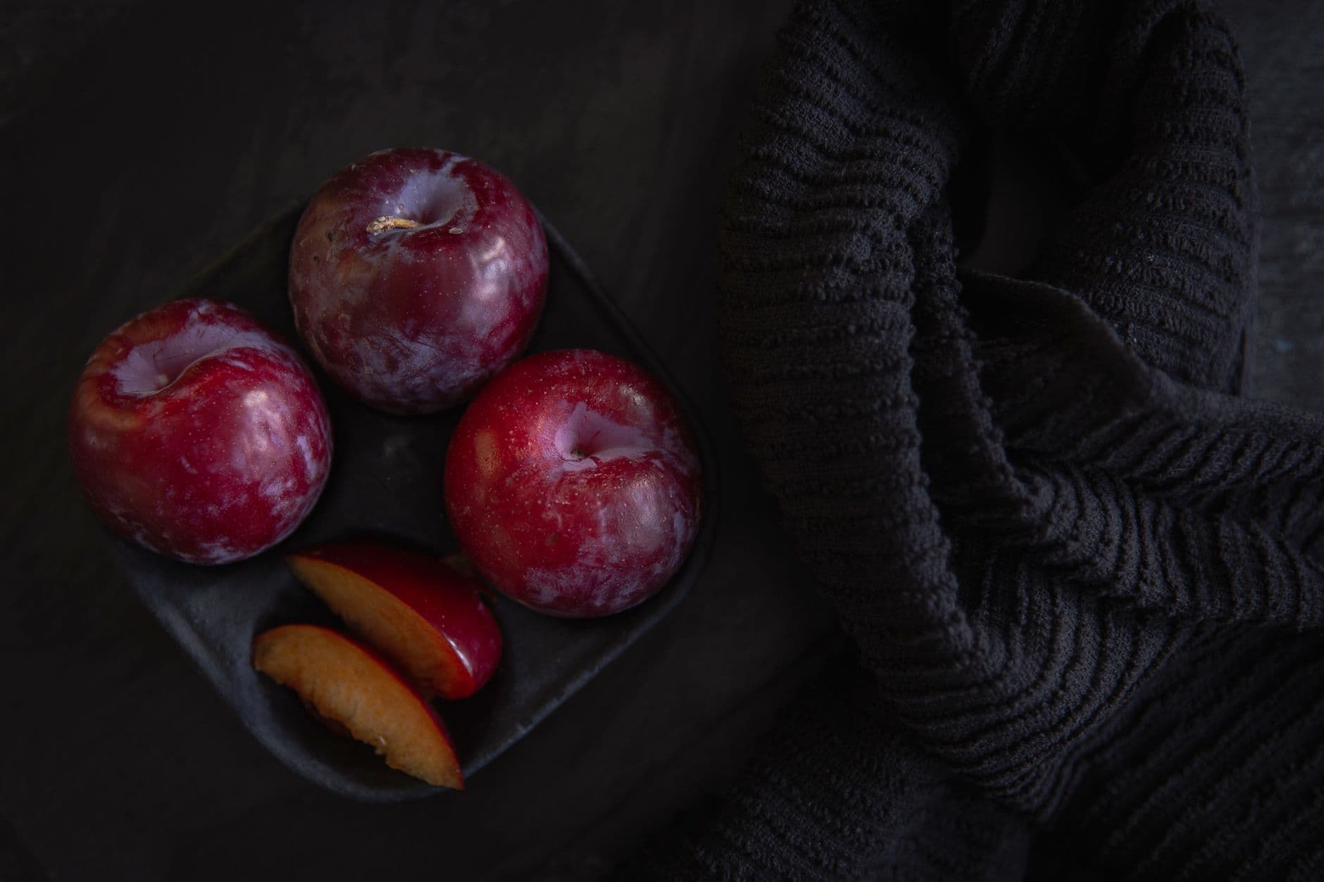 Top view of fresh red plums on a dark textured surface with a black knitted scarf adding contrast.