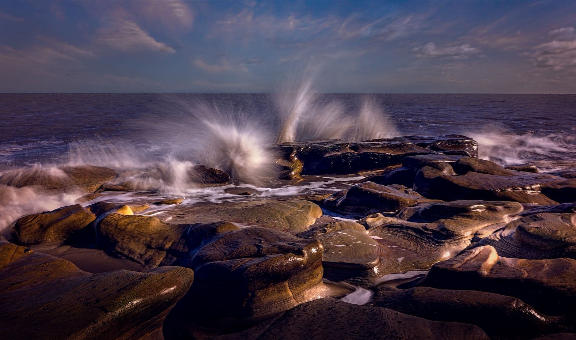 A powerful wave crashes onto a rugged rock formation by the sea, captured during the day.