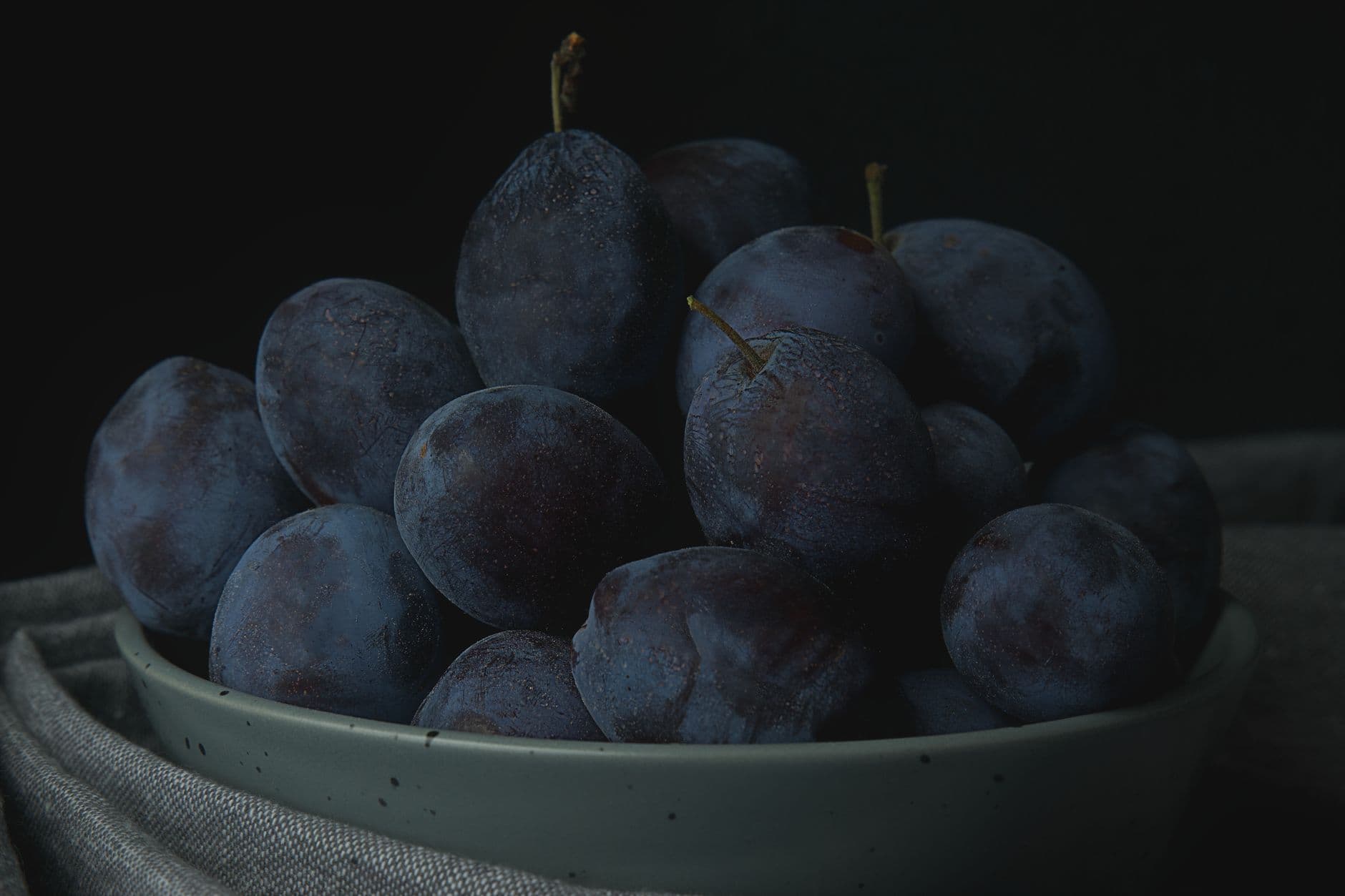 A close-up photo of a bowl filled with fresh, ripe purple plums on a black background.